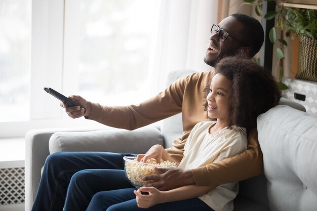 Photo smiling african american young dad relax on couch with cute preschooler daughter watching tv together happy black father sit rest on sofa with girl child use remote enjoy movie time with popcorn