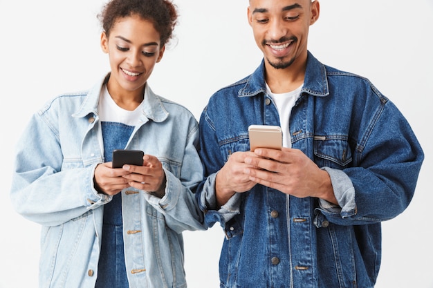 Smiling african couple in denim shirts writing messages on their smartphones over grey wall