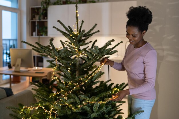 Smiling african female stringing led garland along branches of live christmas tree at cozy home