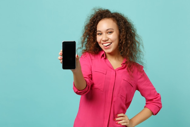 Smiling african girl in casual clothes hold mobile phone with blank empty screen isolated on blue turquoise wall background in studio. People sincere emotions, lifestyle concept. Mock up copy space.