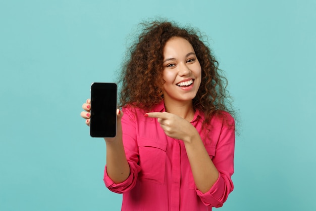 Smiling african girl in casual clothes point index finger on mobile phone with blank empty screen isolated on blue turquoise background. People sincere emotions, lifestyle concept. Mock up copy space.