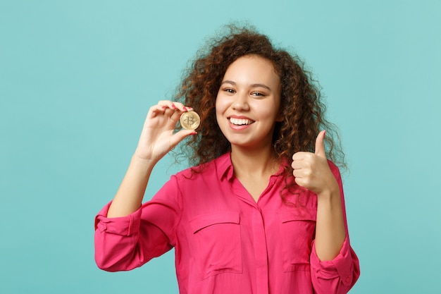 Smiling african girl in casual clothes showing thumb up, holding bitcoin future currency isolated on blue turquoise background in studio. People sincere emotions lifestyle concept. Mock up copy space.