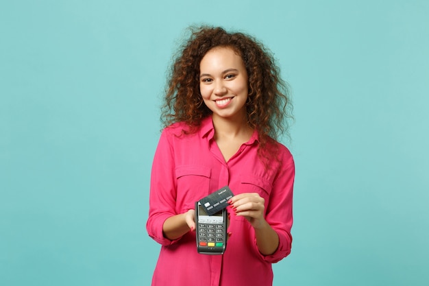 Smiling african girl hold wireless modern bank payment terminal to process, acquire credit card payments isolated on blue turquoise background. People emotions, lifestyle concept. Mock up copy space.