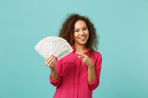 Smiling african girl pointing index finger on fan of money in dollar banknotes, cash money isolated on blue turquoise wall background. People sincere emotions, lifestyle concept. Mock up copy space.