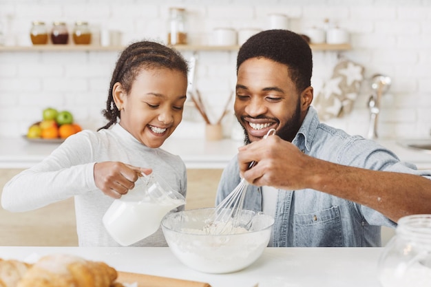 Smiling african girl pouring milk into dough bowl