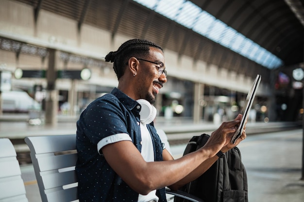 Smiling african male using a tablet computer while he is waiting on a bench at a train station