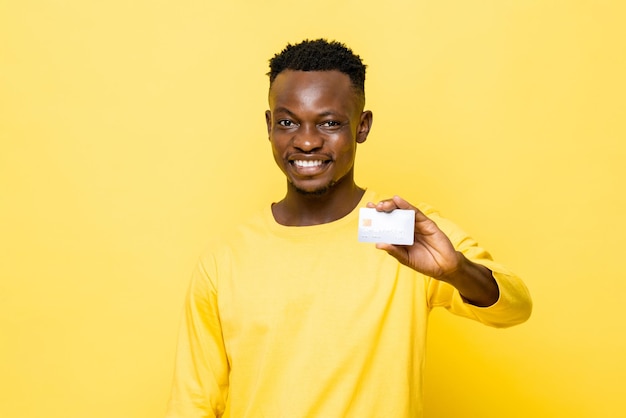 Smiling African man in casual attire holding credit card on isolated yellow studio background