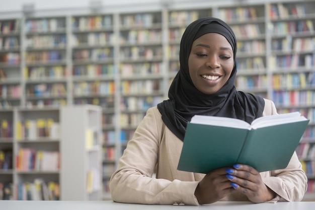 Studente africano sorridente che indossa l'hijab nero che studia il libro di lettura che si siede nella biblioteca moderna