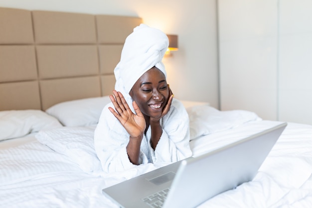 Smiling african woman lying on bed in bathrobe with laptop talking to her friends via video call.