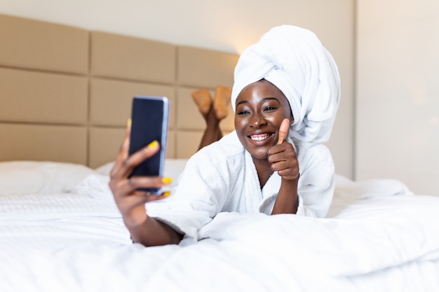 Smiling african woman lying on bed in bathrobe with mobile phone taking a selfie. showing thumbs up