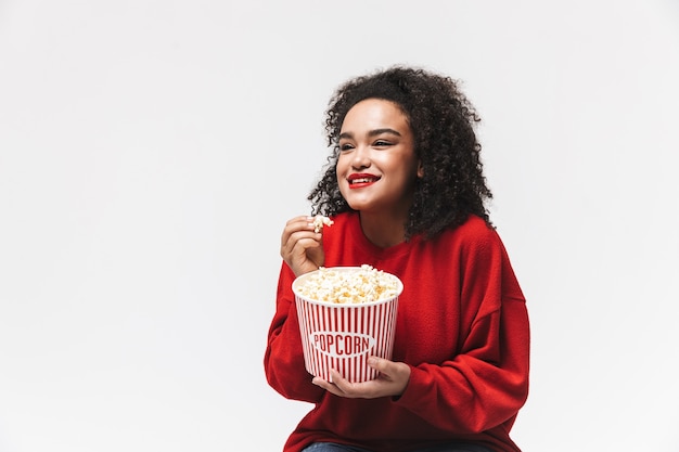 Улыбающаяся африканская женщина в красном свитере ест попкорн и смотрит в сторону на сером фоне