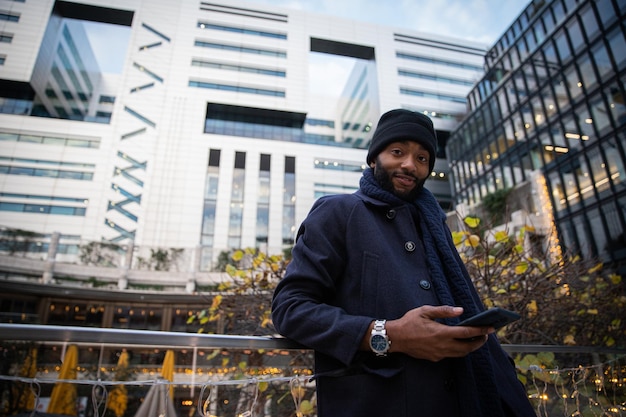 Smiling Afro American businessman elegantly dressed in a black coat and wears a cap during a winter day, he holds his phone in his hand, and wears a watch. Successful young entrepreneur in a big city