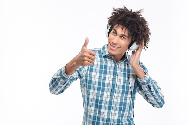 Smiling afro american man listening music in headphones and showing thumb up isolated on a white wall