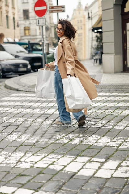 Smiling afro american woman after shopping with paper bags crosses road and looking at camera