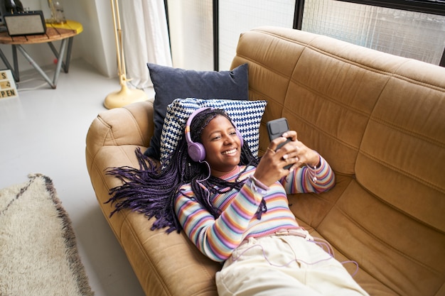 Smiling afro american woman listening to music with headphones and using smartphone relaxing on the ...