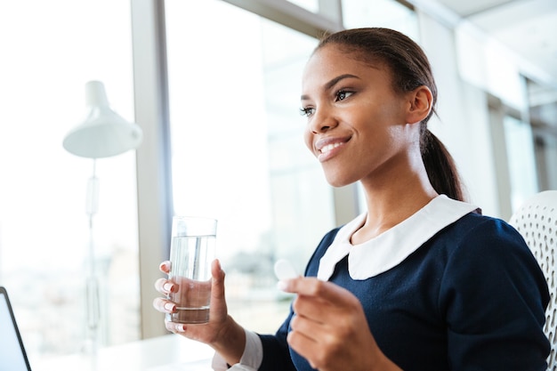 Smiling afro business woman in dress standing near the window and holding water and pill in hands