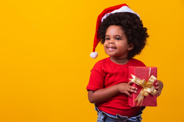 クリスマスプレゼントを手に持ってサンタクロースの赤い帽子でアフロの子供を笑っています。クリスマスのコンセプト。