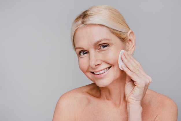 Smiling aged charming woman wiping her face with cotton pod\
isolated over grey background