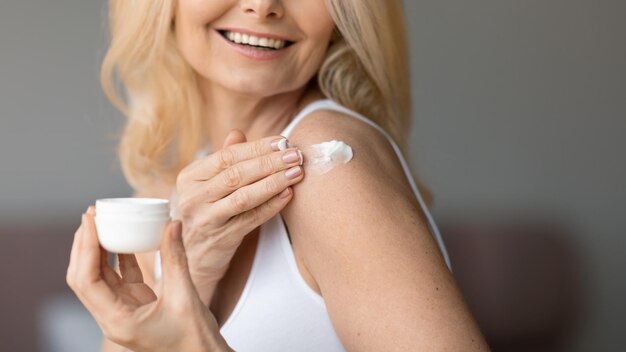 Photo smiling aged female applying cream on shoulder enjoying good morning and daily routine procedures