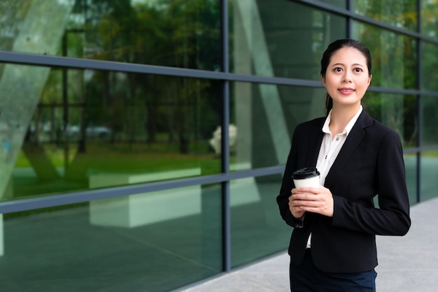 smiling agent rights woman right holding hot espresso cup standing on outside hallway with modern building office transparent windows background in free time.