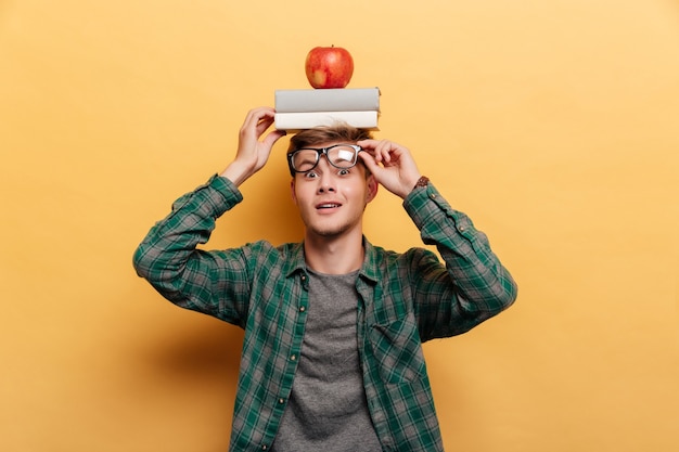 Smiling amazed young man in glasses with book and apple on his head over yellow background