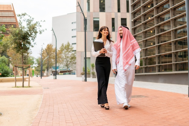 Sorridente uomo d'affari arabo che cammina per la strada della città con la sua giovane e attraente assistente femminile europea