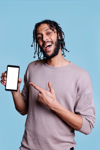 Smiling arab man presenting mobile phone with white screen for application advertisement and looking at camera. Cheerful advertiser pointing with finger at smartphone with empty display for text