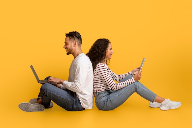 Smiling arab man and woman with different gadgets sitting over yellow background