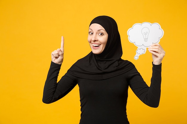 Smiling arabian muslim woman in hijab black clothes hold in hands say cloud with lightbulb isolated on yellow wall  portrait. People religious lifestyle concept. .