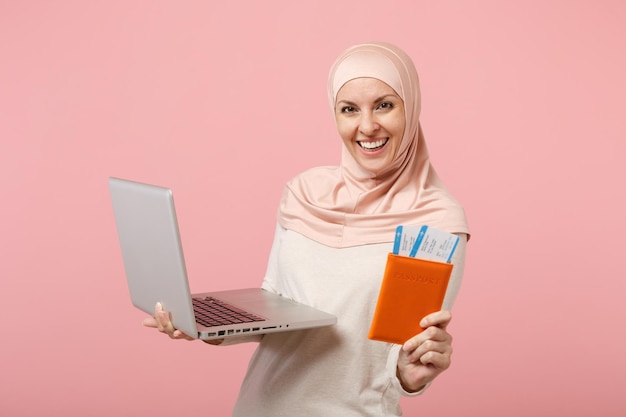 Smiling arabian muslim woman in hijab light clothes posing isolated on pink background. People religious Islam lifestyle concept. Mock up copy space. Holding laptop pc computer, passport and ticket.