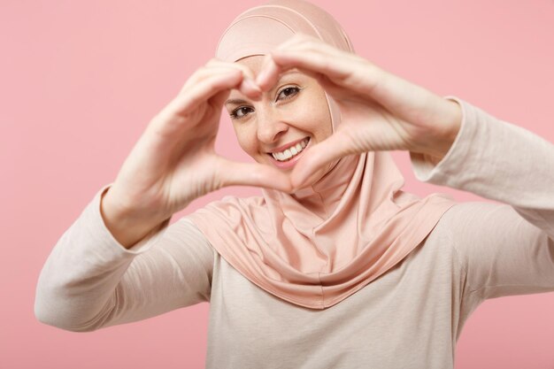 Smiling arabian muslim woman in hijab light clothes posing isolated on pink background. People religious Islam lifestyle concept. Mock up copy space. Showing shape heart with hands, heart-shape sign.