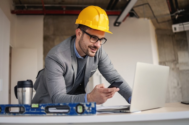 Smiling architect leaning on desk at construction site, looking at laptop and using phone to tell colleagues about project.