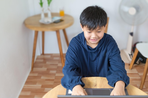 Улыбающийся азиатский мальчик болтает с друзьями или изучает интернет на ноутбуке в социальных сетях дома