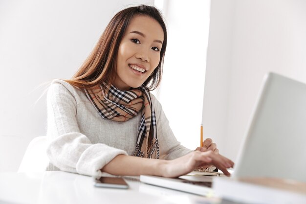 Smiling asian business woman typing something in laptop computer and looking at the front while sitting by the table in office