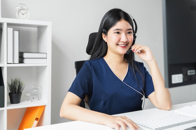 Smiling Asian business woman working in call center at office desk with headset and computer.