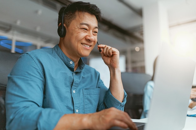 Uomo d'affari asiatico sorridente in cuffia che parla online con il cliente seduto in ufficio