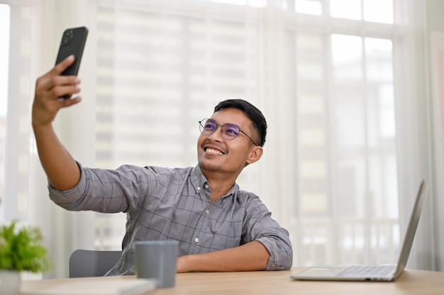 Улыбающийся азиатский бизнесмен делает селфи со своим телефоном, сидя за рабочим столом