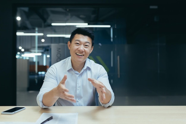 Smiling asian businessman talking on video call looking at camera while sitting at desk in office