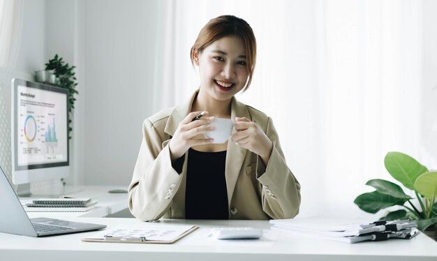 미소 짓는 아시아 사업가 여성이 사무실에서 커피 컵과 노트북을 들고 카메라를 쳐다보고 있습니다.