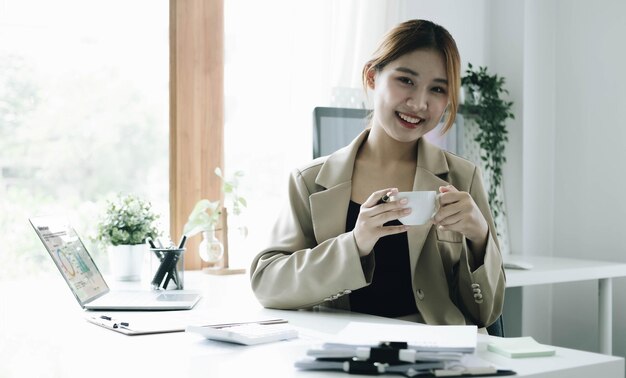 미소 짓는 아시아 사업가 여성이 사무실에서 커피 컵과 노트북을 들고 카메라를 쳐다보고 있습니다.
