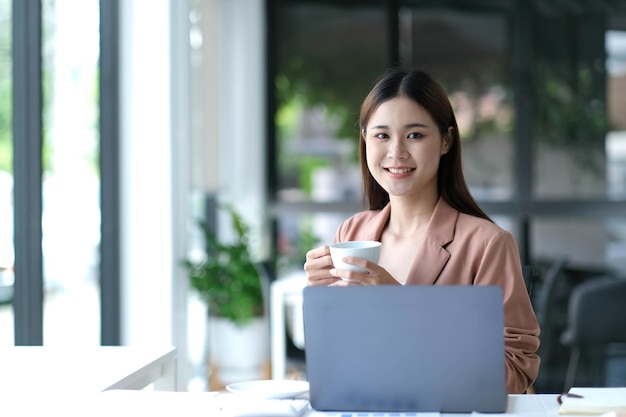 Smiling Asian businesswoman holding a coffee mug and laptop at the office Looking at the camera