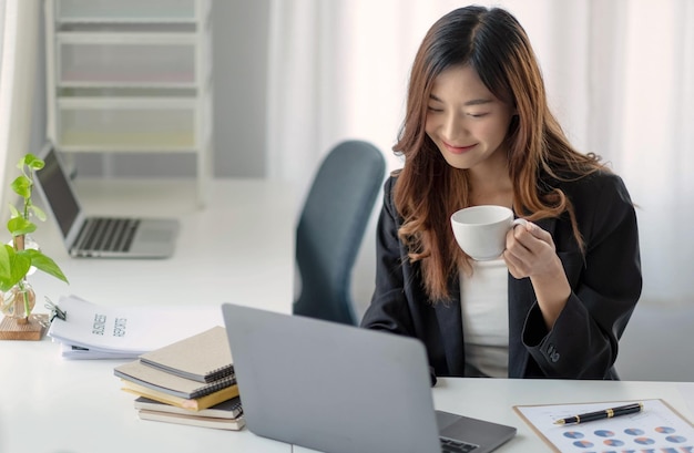 Улыбающаяся азиатская деловая женщина с кружкой кофе и ноутбуком в офисе