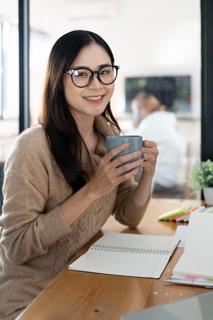 カメラを見てオフィスの机に座ってコーヒーのマグカップを保持している笑顔のアジアの実業家