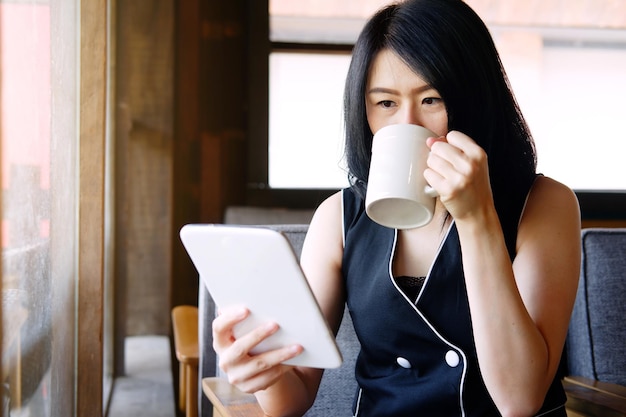 La donna di affari asiatica sorridente sta bevendo una tazza di caffè e sta tenendo il tablet per lo shopping online
