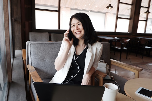 Smiling asian businesswoman is is calling with smartphone online working in social media at workplace in modern office or relaxing at coffee shop. lifstyle of people with techonology concept.