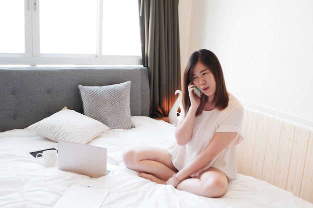 웃고 있는 아시아 여성 사업가가 휴일에 침실에서 스마트폰으로 이야기하고 있습니다. 그녀는 Covod-19 발병 개념 동안 집에서 기술로 온라인 작업을 하고 있습니다.