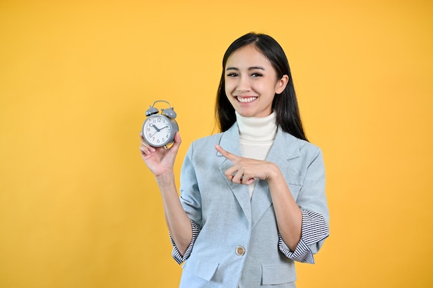 A smiling Asian businesswoman pointing at an alarm clock on her hand isolated yellow background