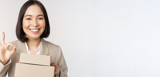 Smiling asian businesswoman showing okay sign and boxes with delivery goods prepare order for client standing over white background