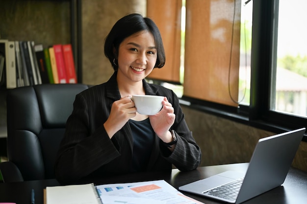 Smiling Asian businesswoman sits at her desk with a coffee cup in her hands