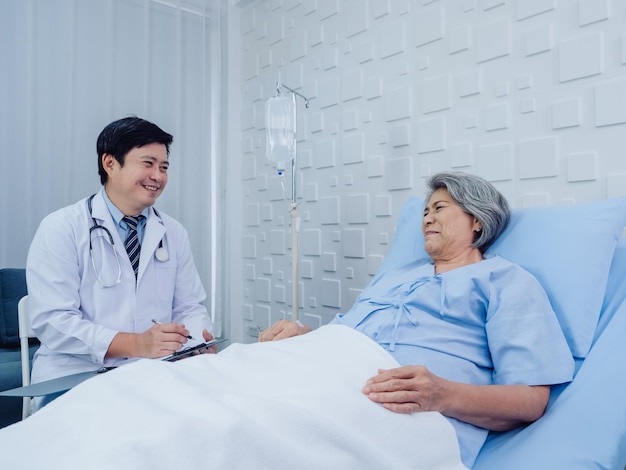 Smiling asian doctor in white suit take notes while discussing\
happy asian elderly senior woman patient who lying on bed with\
receiving saline solution in hospital health care and medical\
concept